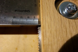 Teeth of the saw blade projecting a few millimeters beyond the depth stop, with a string action gauge held to the wood, showing the depth setting of the set-up (about 2,5 mm)